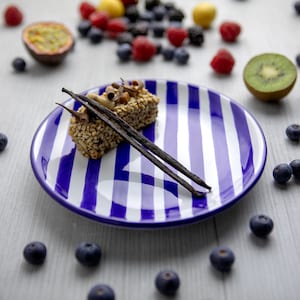 Puede incluir: Un plato de cerámica rayado azul y blanco con un pequeño postre rectangular cubierto con vainas de vainilla. El postre está sobre un fondo blanco con bayas dispersas.