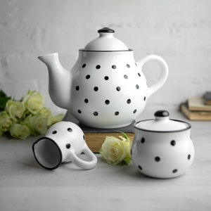 May include: A white teapot, creamer and sugar bowl with black polka dots. The teapot has a black lid and a spout. The creamer and sugar bowl have black lids. The set is on a white surface with a book and flowers.