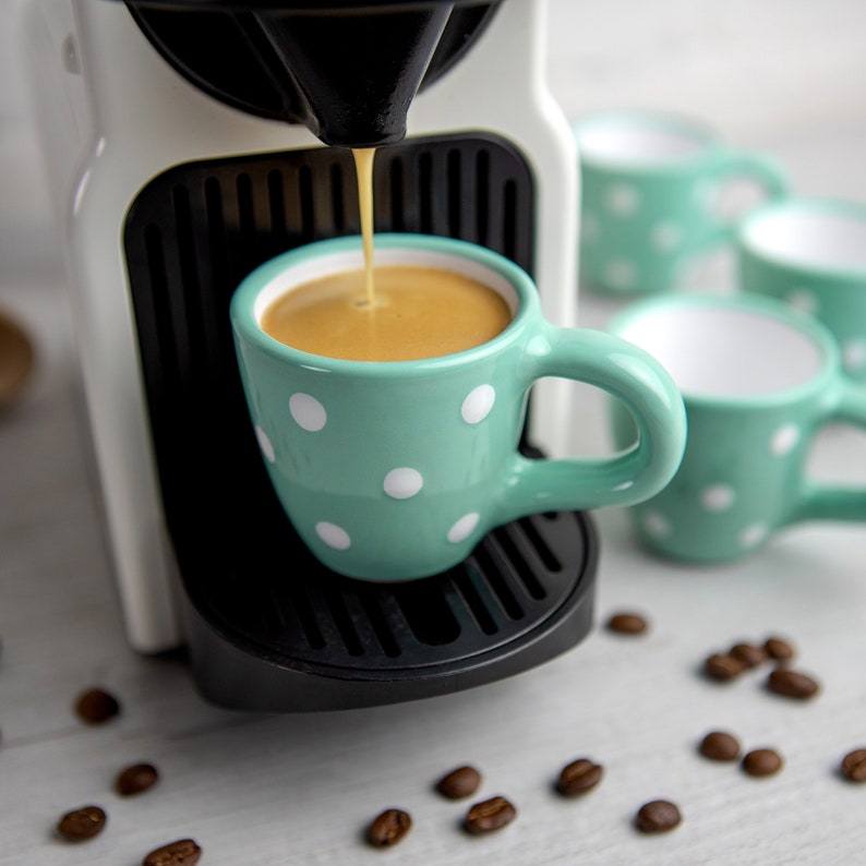 May include: A white espresso machine with a turquoise cup with white polka dots being filled with coffee. The cup is sitting on a black drip tray. There are several turquoise cups with white polka dots on a white surface with coffee beans scattered around.