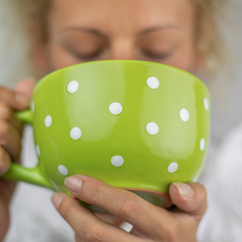 May include: A green ceramic mug with white polka dots. The mug is being held by a person's hands.