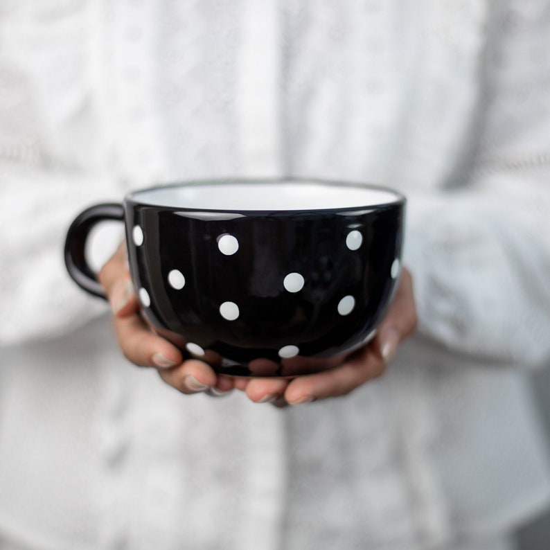 K&ouml;nnte beinhalten: Eine gro&szlig;e Tasse mit schwarzen und wei&szlig;en Punkten und wei&szlig;em Innenraum. Die Tasse wird von den H&auml;nden einer Person gehalten.