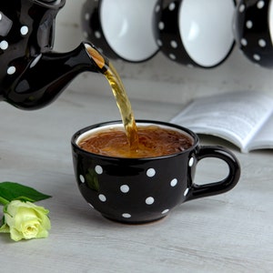K&ouml;nnte beinhalten: Eine schwarze Teekanne mit wei&szlig;en Punkten gie&szlig;t Tee in eine schwarze Teetasse mit wei&szlig;en Punkten. Die Teetasse steht auf einer wei&szlig;en Holzoberfl&auml;che.