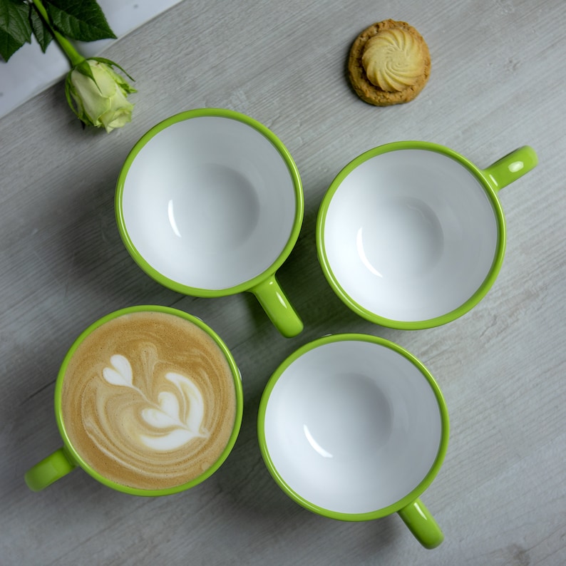 May include: Four green and white mugs, three of which are empty, and one filled with a latte. The latte has a heart-shaped design in the foam. A cookie sits on a wooden surface.