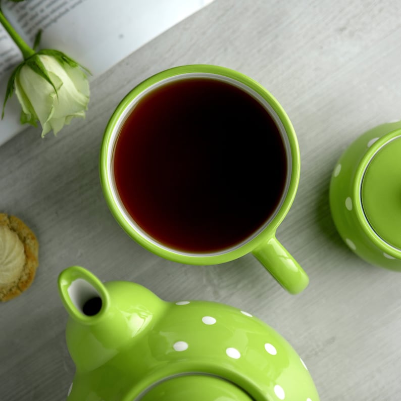 May include: A green teapot with white polka dots, a matching teacup filled with black tea, and a sugar bowl with a lid. The teapot has a spout and a handle. The teacup has a handle. The sugar bowl has a lid and a handle.