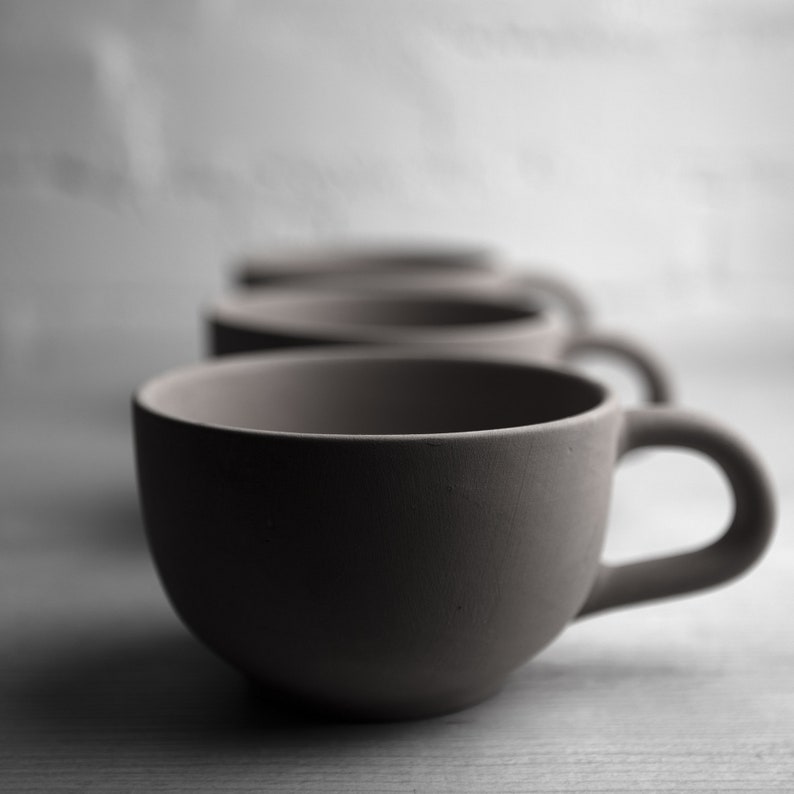 May include: Three ceramic mugs in a row, with the closest mug in focus. The mugs are a light brown color and have a smooth, matte finish.