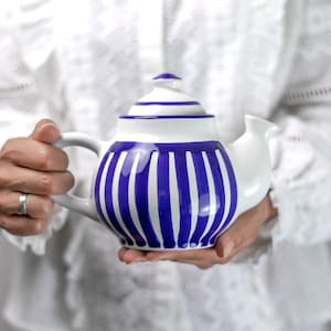 Op de afbeelding: Witte keramische theepot met een blauw gestreept patroon. De theepot heeft een deksel en een tuit. De handgreep is wit. De theepot wordt vastgehouden door een persoon die een wit overhemd draagt.