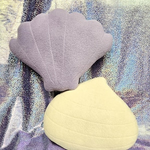 May include: Two seashell-shaped pillows. One is a light lavender color, shaped like a scallop shell. The other is a cream color, resembling a clam shell. Both pillows are made of a soft, textured fabric.