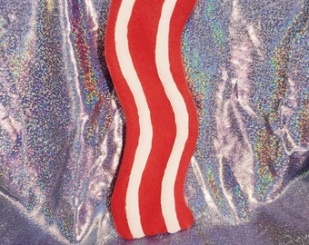 May include: A red and white striped bacon-shaped plush toy. The toy is made of soft, fuzzy material and is perfect for cuddling or playing.