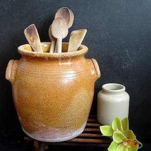 Puede incluir: Un gran tarro de cerámica esmaltada marrón con una boca ancha, lleno de cucharas y espátulas de madera. El tarro tiene un aspecto rústico y vintage y está sentado sobre un soporte de madera.