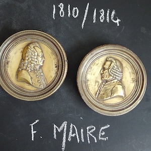 May include: Two antique brass medallions with raised portraits of men in profile. The medallions are framed with a decorative border and have the inscription "F. MAIRE" and the dates "1810/1814" written in white chalk on a black background.