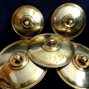Peut inclure: Cinq cymbales en laiton de tailles différentes, empilées les unes sur les autres. Les cymbales sont brillantes et ont une texture légèrement martelée.