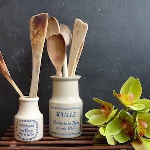 Puede incluir: Dos tarros de cerámica con etiquetas azules y blancas, uno etiquetado "Maille Mostaza de Dijon con vino blanco" y el otro etiquetado "Mostaza de Charroux, Dép. de Palluier". Los tarros están llenos de cucharas de madera.