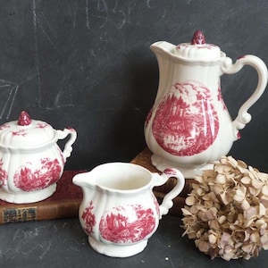 May include: A white ceramic tea set with a red floral pattern. The set includes a teapot, creamer, and sugar bowl.