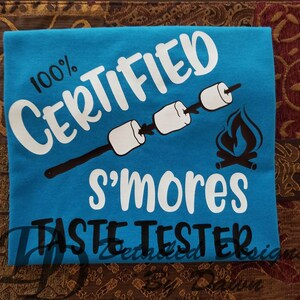 May include: A turquoise t-shirt with white text that reads "100% CERTIFIED S'MORES TASTE TESTER." The design includes illustrations of marshmallows on sticks and a campfire. The shirt is a casual wear item.