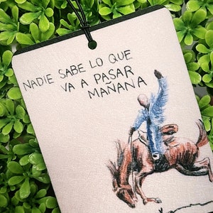 May include: Rectangular air freshener with a black border and a drawing of a cowboy on a bucking bronco. The text "NADIE SABE LO QUE VA A PASAR MAÑANA" is written in black ink. The air freshener hangs from a black string.