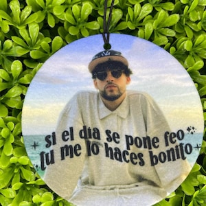 May include: Round air freshener featuring a portrait of a person wearing sunglasses and a baseball cap, with text in Spanish. The air freshener has a black string for hanging and is set against a green, leafy background.