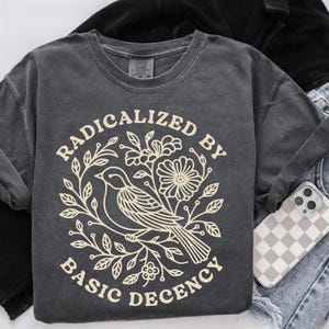 Puede incluir: Sudadera gris oscuro con un diseño crema. El diseño muestra un pájaro en una rama rodeado de flores y hojas, con las palabras "RADICALIZED BY BASIC DECENCY" en un círculo.