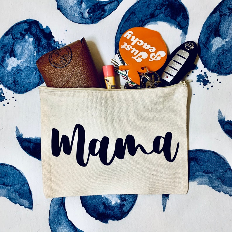 May include: A white canvas zippered pouch with the word "mama" printed in black. The pouch is filled with various items, including a brown leather wallet, a pink lip balm, a key chain with a car key and a keychain charm that says "Just Peachy".