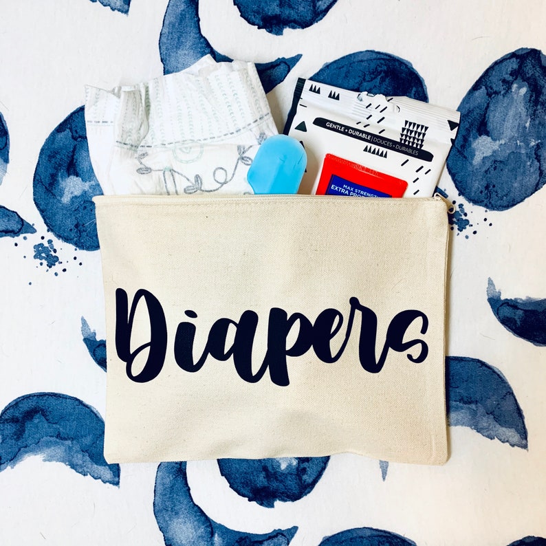 May include: A white canvas zippered pouch with the word "Diapers" printed in black. The pouch is filled with baby supplies, including a diaper, wipes, and a diaper rash cream.