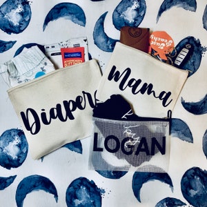 May include: Three canvas zippered pouches with black text. The pouches are labeled "Diaper", "Mama", and "Logan".