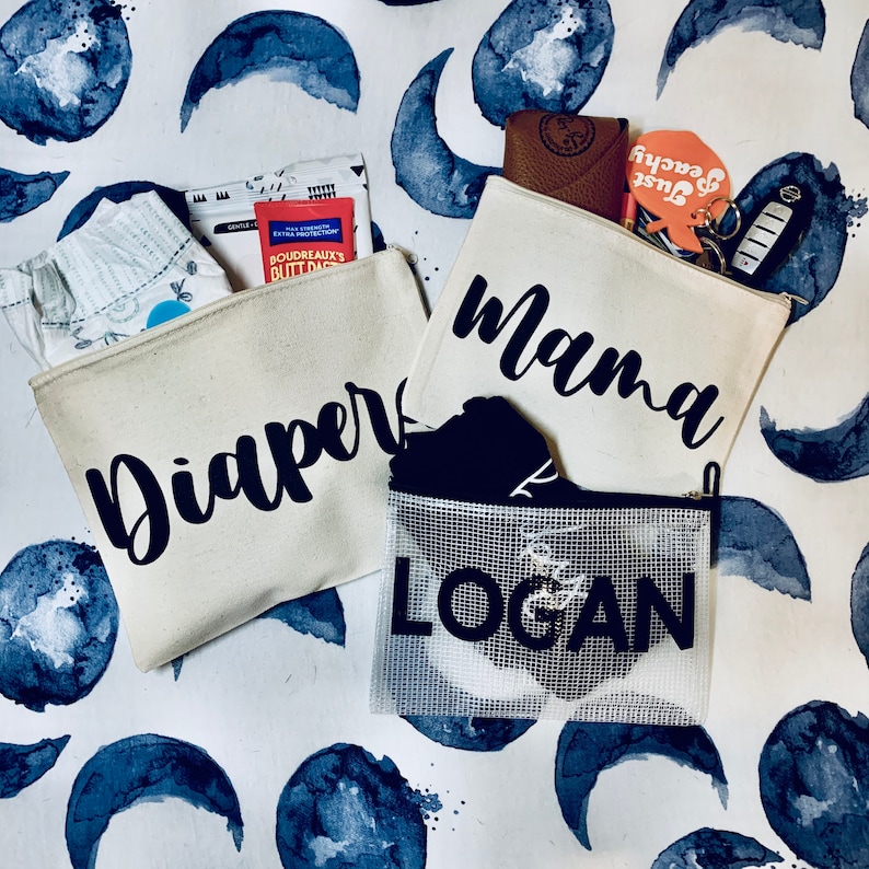 May include: Three canvas zippered pouches with black text. The pouches are labeled "Diaper", "Mama", and "Logan".