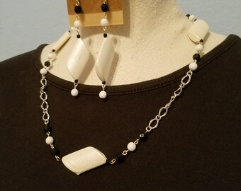 May include: A white and black beaded necklace with a silver chain and white, diamond-shaped pendant. Matching earrings with black beads and white, diamond-shaped pendants.