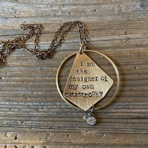 May include: A gold necklace with a pendant that says "I am the designer of my own catastrophe". The pendant is a diamond shape with a circle around it. There is a small crystal hanging from the bottom of the pendant.