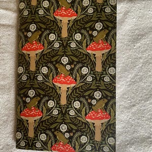 Peut inclure: Un carnet avec un motif répété de champignons rouges et blancs, chacun surmonté d'une grenouille verte, sur un fond sombre avec des accents floraux et de feuilles blanches.