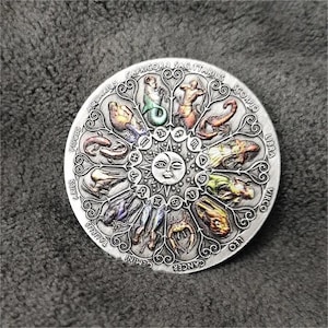 May include: A silver circular zodiac wheel with a sun and moon in the center. The wheel is surrounded by twelve zodiac signs, each with a unique illustration and name. The signs are: Aquarius, Pisces, Aries, Taurus, Gemini, Cancer, Leo, Virgo, Libra, Scorpio, Sagittarius, and Capricorn.