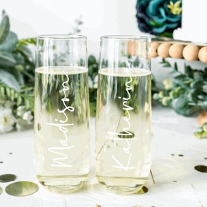 May include: Two clear glass champagne flutes with the names "Madison" and "Katherine" etched on them. The flutes are filled with a clear liquid and are sitting on a white surface with gold confetti.