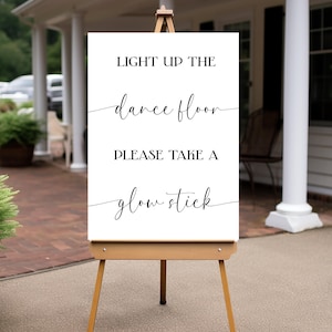 Puede incluir: Un letrero blanco sobre un caballete de madera con el texto "LIGHT UP THE dance floor PLEASE TAKE A glow stick" en escritura negra. El letrero está al aire libre, con un edificio y vegetación en el fondo.