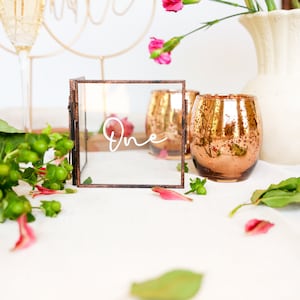 May include: A copper-toned metal frame with a glass insert, displaying the word "One" in white calligraphy. The frame is surrounded by pink flowers, green leaves, and two copper-toned candle holders.