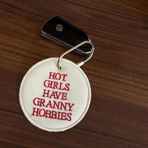 May include: A black car key with a gold keyring attached to a white circular keychain. The keychain has red embroidered text that reads "HOT GIRLS HAVE GRANNY HOBBIES". The key and keychain are on a wooden surface.