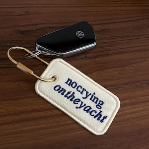 May include: A black car key and a cream-colored keychain with the embroidered phrase "no crying on theyacht" in navy blue. The keychain has a gold-colored metal clasp and ring.
