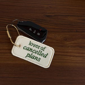May include: Black car key with a gold keychain attached to a cream-colored tag. The tag has the phrase "lover of cancelled plans" embroidered in green. The key and tag rest on a dark wood surface.