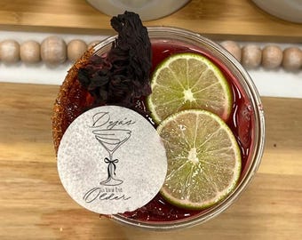 May include: A close-up of a red cocktail with lime slices and a hibiscus flower. The drink is garnished with a sugar rim and a white paper circle with the text "Deja's a tiny bit Older" and a martini glass illustration.