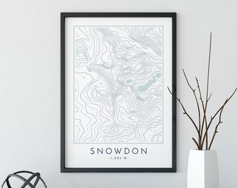 May include: A black framed print with a topographic map of Snowdon, a mountain in Wales. The map is in shades of blue and white, with the name "Snowdon" and the elevation "1,085 M" printed below the map.
