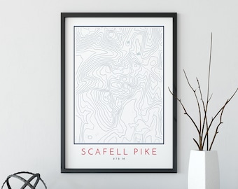 May include: A black framed print with a topographic map of Scafell Pike, a mountain in England. The print has the name "Scafell Pike" and the elevation "978 M" in red text.