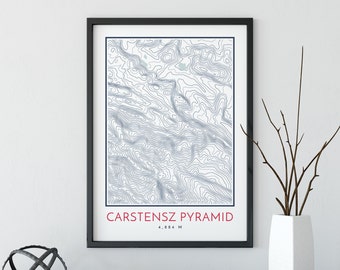 May include: A black framed print with a topographic map of Carstensz Pyramid, a mountain in Indonesia. The print has the text "Carstensz Pyramid 4,884 M" in red at the bottom.