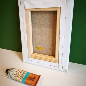 May include: A white canvas stretched over a wooden frame with the text "681581 18X24" printed on the side. A tube of Van Dyke brown oil paint is lying on a white surface.