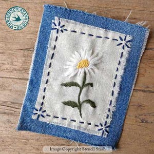 Può includere: Margherita ricamata su un quadrato di tessuto bianco, incorniciata da un bordo in denim blu. La margherita presenta petali bianchi e un centro giallo, con foglie e stelo verdi. Il bordo in denim ha cuciture decorative e bordi sfilacciati. Logo Stencil Stash.
