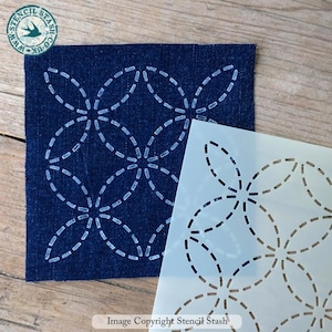 May include: A square, semi-transparent plastic stencil featuring a repeating geometric pattern of interlocking dashed circles. The stencil is positioned next to a piece of dark blue denim fabric that displays the same pattern in white stitching.