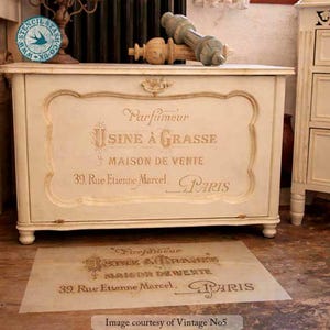 Może przedstawiać: Kremowa, antyczna skrzynia z pozłacanymi detalami i napisem "Parfumeur USINE &Agrave; GRASSE MAISON DE VENTE 39. Rue Etienne Marcel. PARIS." Na podłodze szablon z tym samym tekstem.