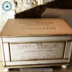 Może przedstawiać: Drewniana skrzynia w stylu vintage pomalowana na kremowo i beżowo. Skrzynia posiada francuski tekst szablonowy, w tym "Esprit de Verveine" i "J. Giraud Fils". Ma prostokątny kształt, pokrywę na zawiasach i spoczywa na małych, zaokrąglonych n&oacute;żkach.