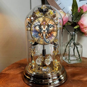 Può includere: Un orologio dorato con un viso di elefante sul quadrante, circondato da cristalli e altri elementi decorativi. L'orologio è racchiuso in una cupola di vetro.