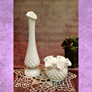 May include: Two white milk glass vases with a textured, dotted surface. The taller vase is a slender bud vase with a flared top. The shorter vase is a bowl-shaped vase with a ruffled edge. Both vases are sitting on a white crocheted doily.