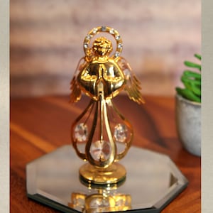 Op de afbeelding: Een gouden engelbeeldje met kristallen accenten staat op een gespiegelde basis. De engel staat in een gebedspositie met zijn handen gevouwen. Het beeldje is een decoratief stuk voor een huis of kantoor.