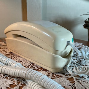 May include: A vintage beige rotary phone with a white coiled cord. The phone is on a white crocheted doily.