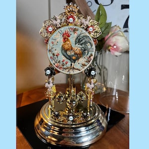 May include: A gold-toned tabletop clock with a rooster design on the face. The clock has a round base and a decorative top with a crown. The clock is surrounded by small, decorative beads.