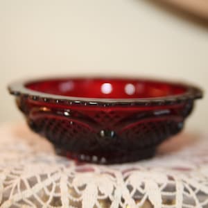 Può includere: Una piccola ciotola di vetro rosso con un motivo decorativo a forma di diamante. La ciotola è appoggiata su un pizzo bianco all'uncinetto.